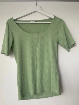 Ribbad topp med knappar - Snygg ljusgrön ribbad topp med korta ärmar och rund halsringning. Toppen har tre dekorativa knappar framtill och är tillverkad i ett mjukt, stretchigt material som sitter skönt på kroppen. Perfekt att styla med jeans eller kjol.