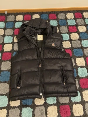 Svart dunväst från Moncler - Snygg svart dunväst från Moncler med huva och två dragkedjeförsedda fickor. Västen har en quiltad design och Moncler-logga på bröstet. Perfekt för lager på lager och ger en sportig vibe. Storlek XL pris går att diskutera
