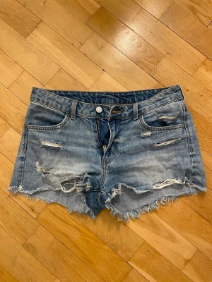 Blå slitna jeansshorts med fransar - Säljer ett par blå jeansshorts med råa fransar och slitna detaljer både fram och bak. Klassisk femficksdesign och knappstängning. Perfekt för dig som gillar en avslappnad och trendig look.