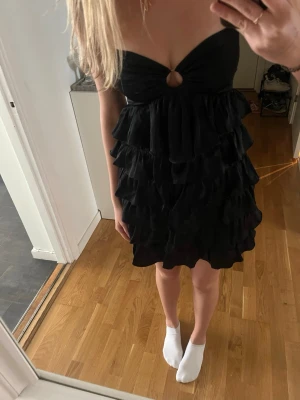 Svart volangklänning - Svart kort klänning med flera lager volanger. Klänningen är axelbandslös och har en liten ringdetalj framtill. Uppsydd efter egna mått men skulle säga strl S.