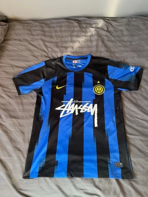 Inter Milan tröja - Säljer en riktigt snygg och unik Inter Milan fotbollströja med det ikoniska Stüssy-trycket på bröstet. Tröjan kombinerar klassiska blåsvarta ränder med en modern "pixlad" design och neongula detaljer. Detaljer: • Skick: Helt ny och oanvänd. • Märke: Nike (Dri-FIT). • Storlek: Large (L). • Design: Inter Milan klubbmärke, Nike swoosh och Stüssy-logga. Storleksreferens: Modellen på bilden är 183 cm lång och väger 72 kg. Som du ser på bilden har den en bra passform – varken för tajt eller för pösig.