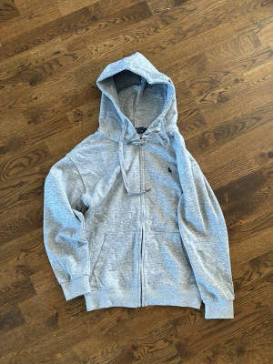 Grå hoodie från Polo Ralph Lauren - Säljer en grå hoodie från Polo Ralph Lauren med dragkedja och huva. Klassisk logga broderad på bröstet. Hoodien har ribbade muddar och fickor framtill. Materialet är mjukt och bekvämt, perfekt för en avslappnad look.