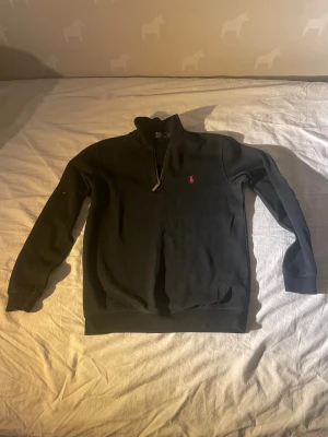 Svart halvzip tröja från Polo Ralph Lauren - Svart långärmad tröja med halvzip från Polo Ralph Lauren. Tröjan har ett litet rött broderat logotyp på bröstet och ribbade muddar vid ärmslut och nederkant. Perfekt för en avslappnad och stilren look.