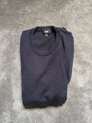 Mörkblå stickad tröja från Uniqlo - Säljer en stilren mörkblå stickad tröja från Uniqlo med rund halsringning och långa ärmar. Tröjan är tillverkad i mjukt material och har ribbstickade muddar vid ärmslut och nederkant. Perfekt för lager-på-lager och enkel att matcha. Har ett hål på en av ärmarna.