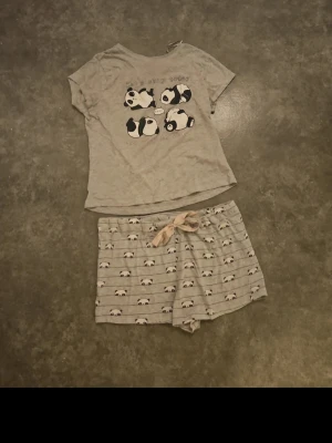 Grå pyjamas med panda-motiv - Pyjamas-set i ljusgrått med kortärmad t-shirt och shorts. T-shirten har söta panda-tryck och texten 'Let's skip today and stay in bed...'. Shortsen har ett mönster med små pandor och en ljus rosett i midjan. Perfekt för mysiga kvällar.