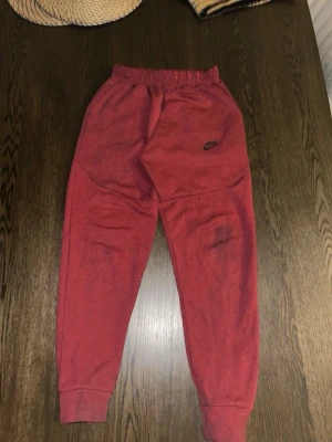 Röda Nike mjukisbyxor med logga - Röda mjukisbyxor från Nike med svart logga på framsidan och elastisk midja. Byxorna har ribbade muddar vid benslut och en svart dragkedja på bakfickan. Perfekta för chill eller träning, tillverkade i mjukt material.