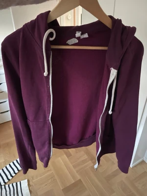 Mörklila hoodie med dragkedja - En mörklila hoodie med vit dragsko och dragkedja framtill. Hoodien har en klassisk huva och två fickor på framsidan. Perfekt för att slänga på sig när det är lite kyligt. Materialet är mjukt och bekvämt, troligen bomullsblandning.