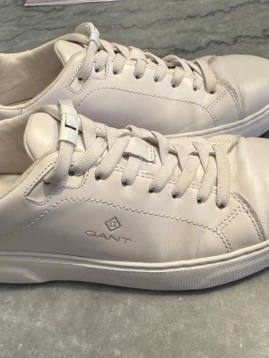 Vita Gant sneakers i läder - Snygga vita sneakers från Gant med klassisk låg profil och stilren design. Skorna är tillverkade i läder och har matchande vita skosnören samt Gant-logga på sidan. Perfekta för dig som gillar en clean och tidlös look.