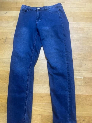 Blå raka jeans med klassisk passform - Säljer ett par blå jeans med raka ben och klassisk femficksdesign. Jeansen har en normal midja och stängs med knapp och dragkedja. Materialet är denim och färgen är mörkblå. Perfekta för dig som gillar en enkel och tidlös stil.
