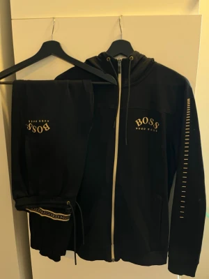 Svart set från Hugo Boss - Snyggt svart set från Hugo Boss med guldfärgade detaljer och logotyp på både tröja och byxa. Setet har dragkedja framtill, huva och ribbade muddar. Materialet är mjukt och bekvämt, perfekt för en cool och avslappnad look.