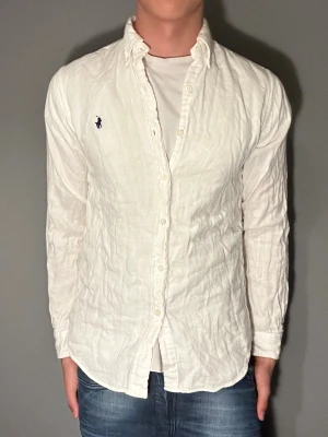 Vit skjorta från Polo Ralph Lauren - Säljer en klassisk vit skjorta från Polo Ralph Lauren med broderad logga på bröstet. Skjortan har lång ärm, knappar framtill och är tillverkad i ett lätt linne-material. Perfekt för dig som gillar stilrena plagg med ikonisk branding.