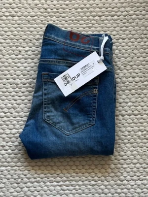 Blå jeans från Dondup - Säljer ett par blå jeans från Dondup med klassisk femficksdesign och snygga slitningar. Jeansen har en rak passform och är tillverkade i denim med en cool tvätt. Perfekta för dig som vill ha ett stilrent och trendigt plagg.