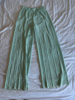 Gröna byxor med ränder - Säljer ett par ljusgröna byxor med vita vertikala ränder. Byxorna har breda ben och elastisk midja för en avslappnad och luftig känsla. Perfekta för sommarens varma dagar och ger en cool vibe till din outfit.