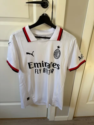 AC Milan vit matchtröja Puma - Säljer en vit AC Milan matchtröja från Puma med svart och röd detalj på krage och ärmslut. Tröjan har klubbens logga, sponsortryck och Puma-logga på bröstet. Materialet är lätt och andas, perfekt för fotboll eller träning.