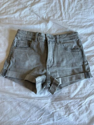 Grå denimshorts med uppvik - Snygga grå denimshorts med uppvikta benslut och klassisk femficksdesign. Shortsen har medel midja, bälteshällor och bakfickor med lock och knapp. Perfekta för varma dagar och enkel att matcha med allt.