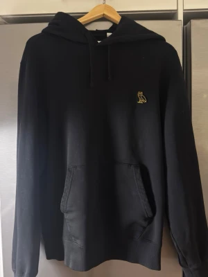 Svart OVO hoodie - Säljer en svart hoodie från OVO med den ikoniska gula uggleloggan broderad på bröstet. Hoodien har huva med dragsko, magficka och långa ärmar. Perfekt för dig som gillar streetwear och vill ha något stilrent och bekvämt.