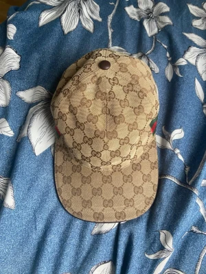Gucci beige keps med GG-mönster - Säljer en beige keps från Gucci med det klassiska GG-monogrammet över hela kepsen. På sidan finns den ikoniska gröna och röda randen. Kepsen har böjd skärm och justerbar rem bak. Materialet är canvas med läderdetaljer.