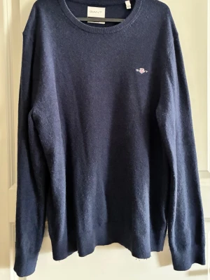 Mörkblå tröja från GANT - Klassisk mörkblå tröja från GANT med rund halsringning och diskret broderad logga på bröstet. Tröjan har långa ärmar och ribbade muddar vid ärmslut och nederkant. Perfekt för en clean och stilren look. L men sitter mer som M