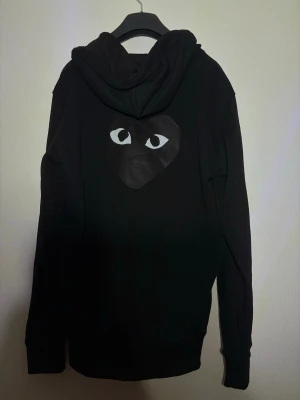 Svart hoodie från Comme des Garçons Play - Säljer en svart hoodie med dragkedja från Comme des Garçons Play. På bröstet och ryggen finns det ikoniska hjärtat med ögon. Hoodien har huva med dragsko och ribbade muddar. Perfekt för dig som gillar streetwear och vill ha något stilrent men ändå unikt.