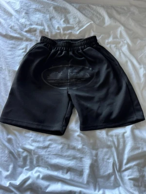 Svarta shorts med skepptryck - Coola svarta shorts med elastisk midja och ett stort tryck av ett skepp på framsidan. Materialet är mjukt och verkar vara polyester, perfekt för en avslappnad och sportig look. Passar dig som vill sticka ut med ett unikt motiv.