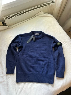 Mörkblå stickad tröja från Stone Island - Snygg mörkblå stickad tröja från Stone Island med klassisk rund halsringning och ribbade muddar. Tröjan har den ikoniska Stone Island-loggan på vänster ärm och är tillverkad i mjukt material som passar perfekt till höst och vinter.