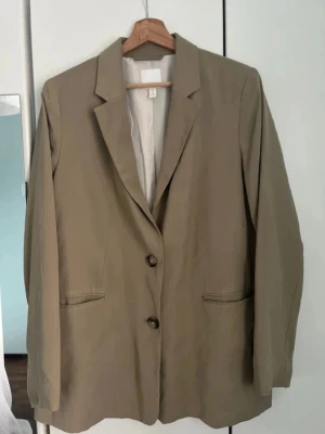 Oversized kavaj - Stilren grön kavaj i oversized modell med två bruna knappar framtill och klassisk krage. Kavajen har två fickor utan lock och är helfodrad. Perfekt för en trendig och avslappnad look.