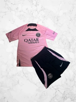 PSG rosa matchställ från Nike - Säljer ett PSG matchställ från Nike med rosa tröja och svarta shorts. Tröjan har PSG-logga, Qatar Airways-tryck och svarta detaljer vid halsen. Shortsen är svarta med rosa inslag och PSG-märke. Perfekt för fotbollsträning eller match. Helt oanvänd