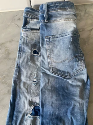 Jack and Jones Jeans  - Säljer ett par riktigt schyssta jeans från Jack & Jones i modellen Glenn. Det är en klassisk slim fit-modell som sitter bra utan att vara för tight. Dessa har en cool "distressed" look med slitningar och hål som är snyggt lagade med mörkare denimtyg bakom, vilket ger dem en unik och rå karaktär. Detaljer: • Modell: Glenn Slim Fit • Märke: Jack & Jones • Storlek: W30 L32 • Färg: Ljusblå tvätt med snygga sli