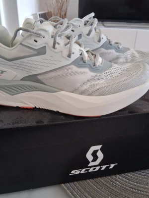 Vita löparskor från Scott - Säljer ett par vita löparskor från Scott med grå detaljer och meshmaterial för bra ventilation. Skorna har tjock sula för optimal dämpning och klassisk snörning. Stl 40 men passar bäst för den med stl 38-39. 