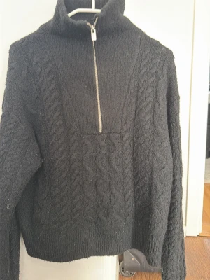 Polo tröja  - Snygg svart stickad tröja med kabelstickat mönster och hög krage. Tröjan har en halv dragkedja framtill och ribbade muddar vid ärmslut och nederkant. Perfekt för kyliga dagar och enkel att matcha till olika outfits.