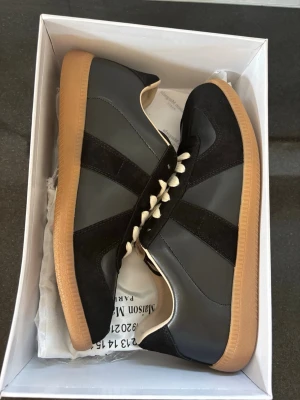 Svarta sneakers med gummisula - Maison Margiela sneakers i svart läder och mocka med klassisk design och beige gummisula. Skorna har vita snören och diskreta detaljer på hälen. Perfekt för dig som gillar stilrena och tidlösa sneakers.