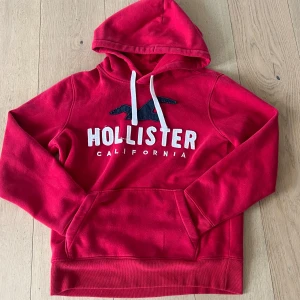 Röd hoodie från Hollister XS - Snygg röd hoodie från Hollister i storlek XS med vit broderad logga och text på bröstet. Tröjan har huva med vita snören och en stor magficka. Perfekt för dig som gillar en avslappnad och sportig stil.