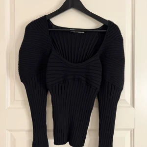 Svart ribbad topp med markerade axlar från NA-KD - Svart ribbad topp med markerade axlar och djup v-ringning. Toppen har lång ärm och en figurnära passform som framhäver siluetten. Användt enstaka gånger. Den har även ett lite tjockare material.
