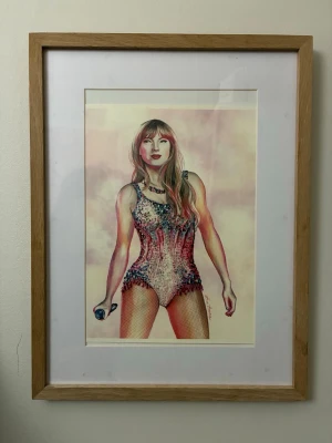 Inramad affisch av Talor Swift   - Snygg affisch med illustration av Talor Swift i scenkläder, inramad med ljus träram och vit passepartout. Motivet visar en person med långt hår, glittrig body och mikrofon i handen mot en pastellfärgad bakgrund. Perfekt som dekoration till rummet. Diskuteras pris