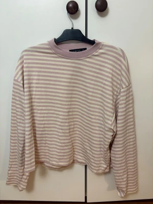 Randig långärmad tröja från Lioness - Säljer en söt långärmad tröja från Lioness i storlek M. Tröjan har ljuslila/rosa och cremevita horisontella ränder, rund halsringning och avslappnad passform. Perfekt för en chill och stilren look. Materialet känns mjukt och bekvämt.