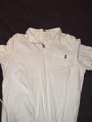 Vit pikétröja från Pier One - Klassisk vit pikétröja från Pier One med korta ärmar och krage. Tröjan har en liten bröstficka med diskret randig detalj i rött och blått samt knappslå framtill. Perfekt till jeans eller shorts för en clean look.