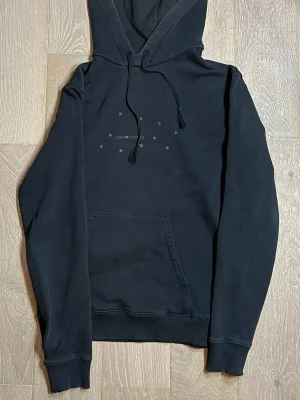 Svart hoodie från Saint Laurent - Svart hoodie från Saint Laurent med dragsko i huvan och känguruficka framtill. Diskret tryck med små stjärnor och texten 'SAINT LAURENT' på bröstet. Tillverkad i mjuk bomull och har ribbade muddar vid ärmar och nederkant. Perfekt för en avslappnad streetwear-look. Storlek S passar även XS, nypris 5300