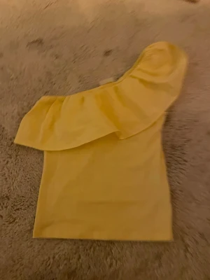 Gul oneshoulder topp från Kappahl - Snygg gul oneshoulder topp från Kappahl med volangdetalj över axeln. Perfekt för dig som vill sticka ut med en färgglad och trendig look. Mjuk och skön att bära, passar till många olika stilar.