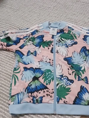 Blommig adidas jacka med fjärilar - Snygg jacka från adidas med blommigt och fjärilsmönster i blått, grönt och vitt på ljusrosa botten. Jackan har ljusblå muddar, dragkedja framtill och klassiska adidas-ränder på ärmarna. Perfekt för dig som vill sticka ut med färg och mönster.