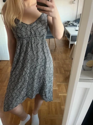 Svartvit mönstrad sommarklänning - Säljer en svartvit klänning med grafiskt mönster och smala axelband. Klänningen har v-ringning och en lätt utställd kjol som ger en avslappnad siluett. Perfekt för varma dagar och enkel att styla med sneakers eller sandaler.
