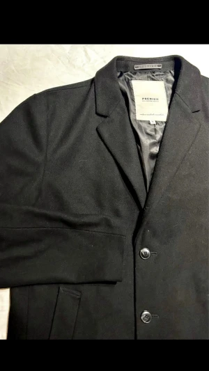 Jack & Jones Premium svart ullkappa XL – stilren rock  - Stilren svart ullkappa från Jack & Jones Premium i strl XL. Klassisk modell som passar både vardag och uppklätt. 50% ull.  Normalt slitage. Mått: Byst ca 59cm, Längd ca 96cm,        Ärm ca 69cm,                                                Axel ca 50cm.