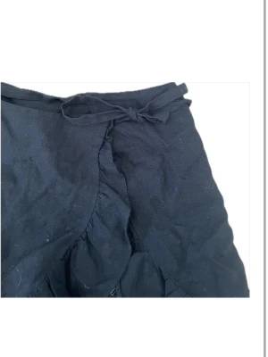 Svart omlottkjol med volang - Snygg svart omlottkjol i 100% linne med volangkant och knytband i midjan. Kjolen har en feminin och trendig look med fina detaljer framtill. Perfekt att matcha med en enkel topp eller stickad tröja. Gjord i ett mjukt tyg som känns bekväm.