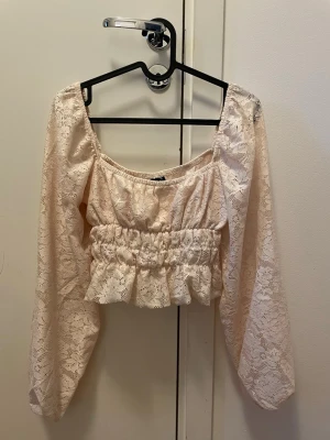 Beige spetsig croppad blus - Söt croppad blus i beige spets med långa ärmar och volang nedtill. Toppen har en smockad midja som ger en snygg passform och romantisk känsla. Perfekt för dig som gillar feminina detaljer och vill sticka ut lite extra.