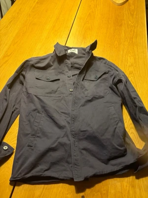 Mörkblå overshirt med fickor - Stilren mörkblå overshirt med två bröstfickor med lock och knapp, samt sidofickor. Långärmad modell med klassisk krage och knäppning framtill. Perfekt att bära som lager över t-shirt. Materialet ser ut att vara bomull eller bomullsblandning. Large men passar M också