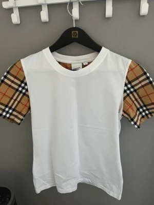 Vit Burberry t-shirt med rutiga ärmar - Snygg vit t-shirt från Burberry med klassiska beige, röda och svarta rutiga ärmar. T-shirten har rund halsringning och är tillverkad i mjukt bomullsmaterial. Perfekt för dig som vill ha en stilren men ändå unik look.