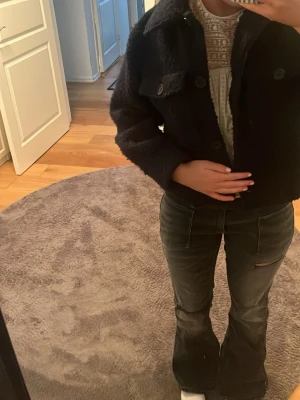 Mörkblå ullig cardiganjacka - Säljer en mörkblå cardiganjacka med ullig textur och stora knappar framtill. Jackan har två bröstfickor med lock och knapp, samt en klassisk krage. Perfekt för dig som vill ha en trendig och mysig jacka till hösten.