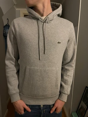 Grå hoodie från Lacoste med logga - Snygg grå hoodie från Lacoste med klassisk krokodillogga på bröstet. Hoodien har justerbar snörning i huvan, stor magficka och ribbade muddar vid ärmar och midja. Tillverkad i mjuk bomull för skön känsla och passar perfekt till jeans. Pris kan diskuteras, hör av er vid frågor! 