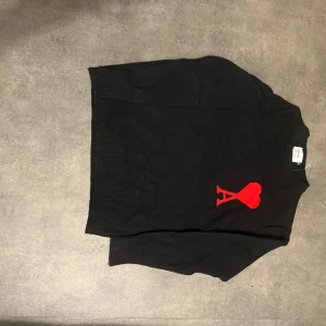 Svart stickad tröja Ami Paris  - Svart stickad tröja från Ami Paris med röd hjärtlogga och bokstaven A på bröstet. Tröjan har rund halsringning, ribbade muddar och lång ärm. Perfekt för dig som vill ha en stilren men ändå unik look.