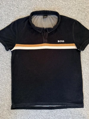 Svart topp från BOSS - Svart kortärmad topp från BOSS med vit och beige rand över bröstet. Gjord i mjuk material. Ca 67cm på längden och 52 cm på brädden. Hör gärna av dig vid funderingar och behov av fler bilder!🤗