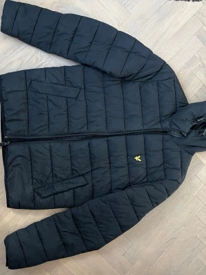 Svart pufferjacka från Lyle & Scott - Snygg svart pufferjacka från Lyle & Scott med gul logga på bröstet. Jackan har raka kanaler, två stora fickor framtill och dragkedja hela vägen upp. Perfekt för kalla dagar och har en klassisk, stilren look.